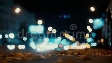 城市灯光和汽车在交通背景下行驶。 秋天的树叶从路过的汽车上随风飘散在夜路上。